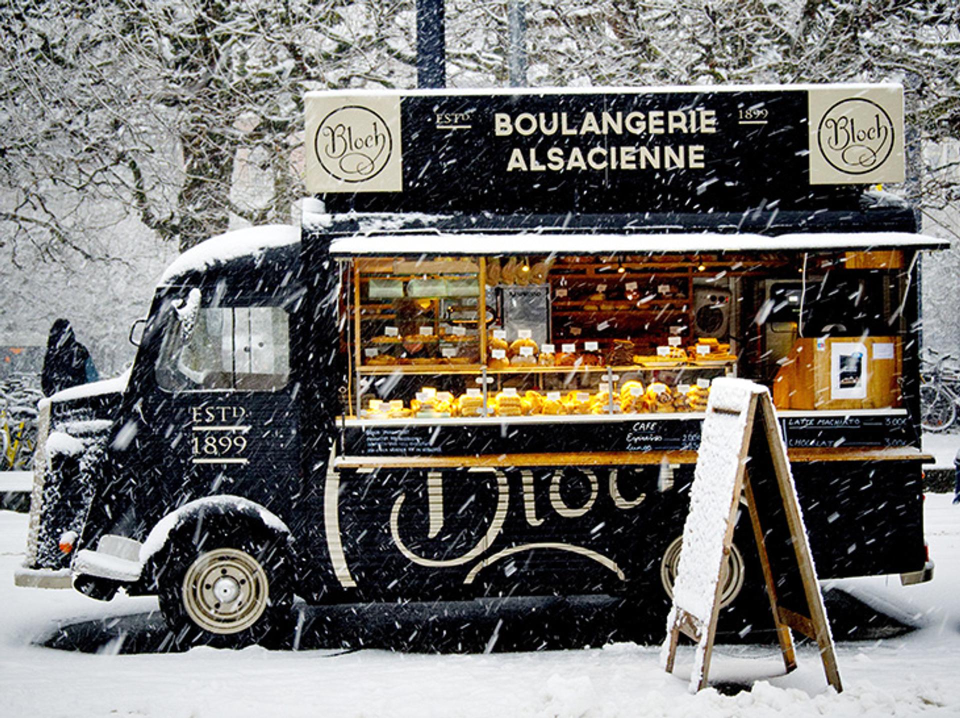 camión de comida Alsacience Boulangerie negro estacionado en medio de una tormenta de nieve y con un símbolo fuera